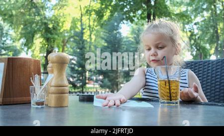 Odessa, Ukraine, Europe de l'est. 3rd juillet 2022. La petite fille est assise dans un café. Blonde fille examine sa main tout en étant assise dans un café de rue sur le parkk. (Credit image: © Andrey Nekrasov/ZUMA Press Wire) Banque D'Images