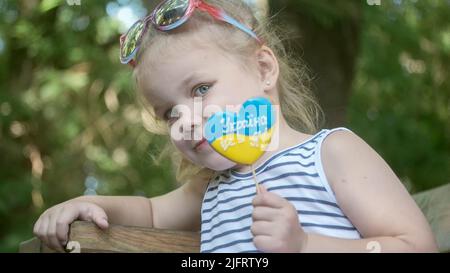 3 juillet 2022, Odessa, Ukraine, Europe de l'est: Une petite fille blonde tient du pain d'épice dans les couleurs nationales ukrainiennes dans sa main, il dit ''l'Ukraine est moi''. Portrait en gros plan d'une fille assise sur le banc du parc avec des biscuits aux couleurs du drapeau ukrainien. Ralenti. Odessa Ukraine (Credit image: © Andrey Nekrasov/ZUMA Press Wire) Banque D'Images