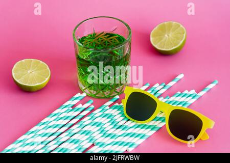 lunettes de soleil et verre d'alcool vert boire près des pailles en papier rayé et des limes sur le rose Banque D'Images