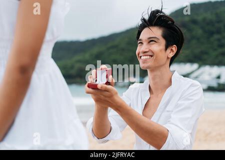 Joyeux souriant asiatique 30s homme proposant une femme avec un anneau de mariage sur la plage en été. Un couple portant une chemise et une robe blanches - objectifs de relation et concept de proposition de mariage Banque D'Images
