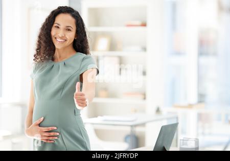 Gérer sa grossesse et sa carrière professionnelle en toute transparence. Portrait d'une femme d'affaires enceinte montrant les pouces dans un bureau. Banque D'Images