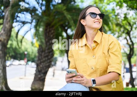 Attrayante femme gaie utilisant un smartphone assis dans le parc, souriante femme étudiant dans des lunettes de soleil tenant le téléphone et regardant de côté, textant en ligne Banque D'Images