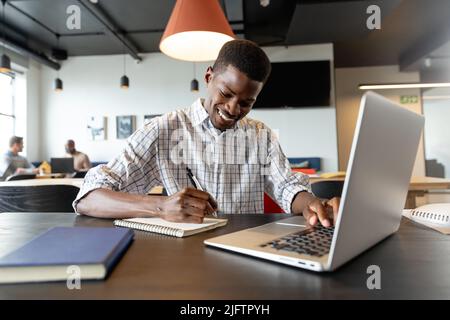 Un jeune homme d'affaires afro-américain souriant écrit dans un journal intime et utilise un ordinateur portable dans un bureau créatif Banque D'Images