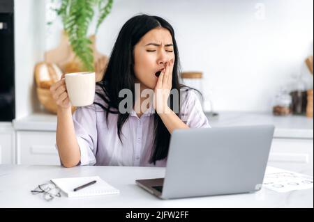 Sleepy Asian jolie fille travaillant à distance, gestionnaire de contenu, indépendant, étudiant, assis à la table dans la cuisine avec un ordinateur portable, le début de la journée de travail, boire du café, bâilloir, incapable de se réveiller Banque D'Images