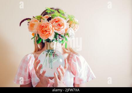 La jeune femme cache le visage derrière un vase avec un bouquet de fleurs. Arrangement avec des roses d'orange. Esthétique dans la décoration d'intérieur Banque D'Images