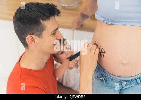 Sourire un jeune papa caucasien dans un t-shirt orange écrit sur le ventre de sa femme enceinte. S'attendre à une autre progéniture. Photo de haute qualité Banque D'Images