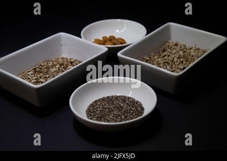 Amandes, chia et autres graines végétaliennes saines dans des bols en porcelaine blanche sur fond noir Banque D'Images