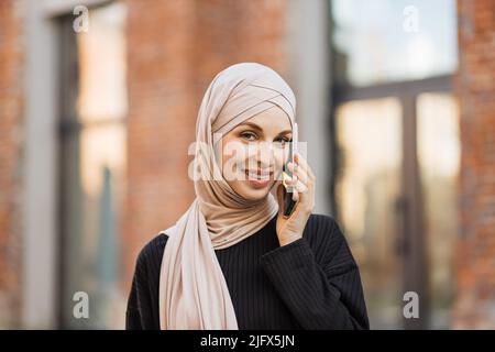 Femme musulmane souriante vêtue d'un costume noir tendance qui marche dans la rue et utilise son téléphone. Jolie femme arabe d'été dans hijab marche dans la rue avec le téléphone mobile. Banque D'Images