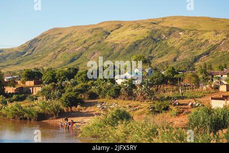 Ihosy, Madagascar - 05 mai 2019: Rivière lente boueuse, herbe verte et buissons sur le côté, petites montagnes en arrière-plan, les femmes laver les vêtements dans l'eau au shor Banque D'Images