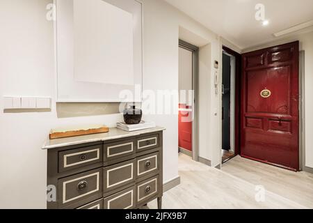 Hall d'entrée et salon avec parquet clair, boiseries blanches, buffets en bois avec tiroirs gris et miroir et porte d'entrée vintage en rouge Banque D'Images