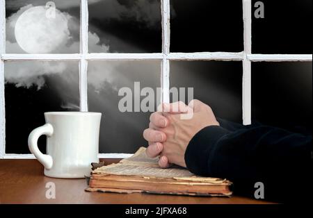 Priant les mains sur une vieille bible ouverte avec une tasse de café blanc et des faisceaux de lune illuminant dans la fenêtre Banque D'Images