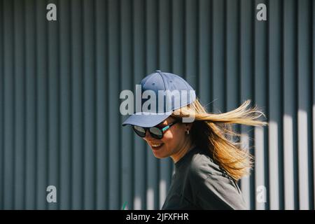 une fille dans une casquette et des lunettes de soleil sourit contre un mur gris nervuré Banque D'Images