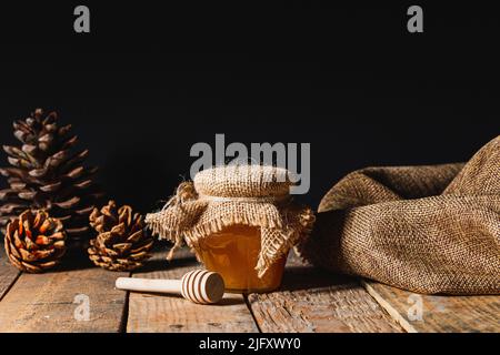 Rustique encore la vie dans laquelle vous pouvez voir un pot en verre avec du miel recouvert d'un tissu de sackCloth, à côté d'elle un bâton de miel en bois et quelques cônes de pin. Everythin Banque D'Images