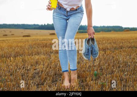 une fille pieds nus avec des baskets et une tasse en carton avec du café à la main debout dans le champ agricole avec des botte de foin et des balles après la récolte. Banque D'Images