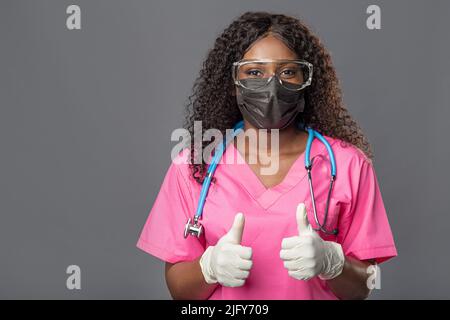 Jeune femme afro-américaine médecin en gants masque et lunettes sur fond gris souriant détendu et gai. Succès et optimisme Banque D'Images