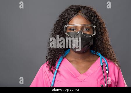 Jeune femme afro-américaine médecin en gants masque et lunettes sur fond gris souriant détendu et gai. Succès et optimisme Banque D'Images