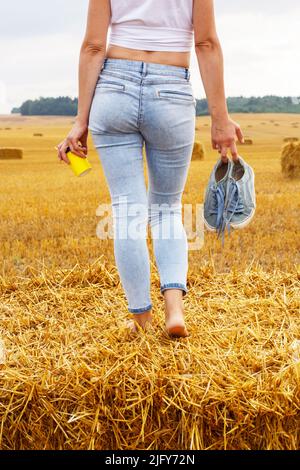 une fille pieds nus avec des baskets et une tasse en carton avec du café à la main debout dans le champ agricole avec des botte de foin et des balles après la récolte. Banque D'Images