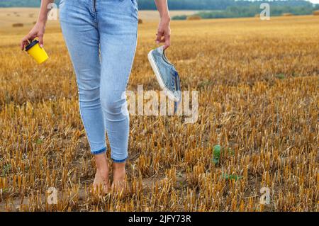 une fille pieds nus avec des baskets et une tasse en carton avec du café à la main debout dans le champ agricole avec des botte de foin et des balles après la récolte. Banque D'Images