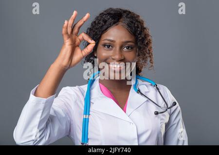 Femme africaine médecin en manteau blanc et avec un stéthoscope montre un signe d'approbation avec ses mains, pouces vers le haut, d'accord. Banque D'Images