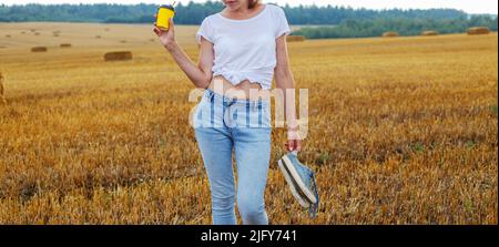 une fille pieds nus avec des baskets et une tasse en carton avec du café à la main debout dans le champ agricole avec des botte de foin et des balles après la récolte. Banque D'Images