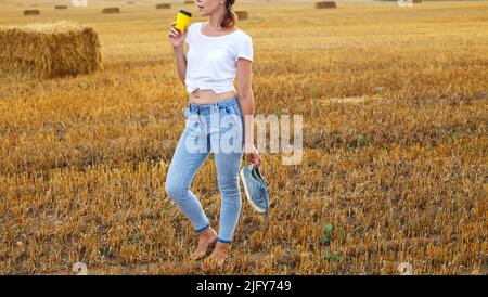 une fille pieds nus avec des baskets et une tasse en carton avec du café à la main debout dans le champ agricole avec des botte de foin et des balles après la récolte. Banque D'Images