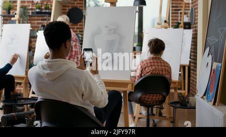 Étudiant créatif prenant la photo d'un croquis de vase sur un smartphone en cours d'art. Jeune homme utilisant le téléphone portable pour prendre des photos et se souvenir de la technique de dessin lors de la leçon éducative. Banque D'Images