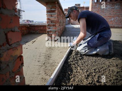 Vue latérale du jeune travailleur dans des gants de protection nivelant la dalle de béton à l'aide de la truelle à l'air frais. Concept de processus de travail avec le ciment et les équipements spéciaux en bon temps. Banque D'Images