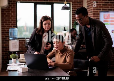 Les employés de l'entreprise font du travail d'équipe dans un bureau adapté aux personnes handicapées, en utilisant un ordinateur portable pour naviguer sur Internet. Employée féminine occupant un fauteuil roulant avec une déficience physique travaillant sur la stratégie du projet. Banque D'Images