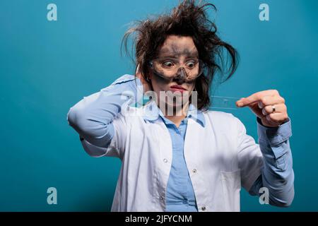 Scientifique fou avec cheveux désordonnés et visage sale regardant confus au plateau de lames de microscope sur fond bleu. Un biochimiste fou et fou qui regarde un échantillon microscopique après une explosion chimique. Banque D'Images