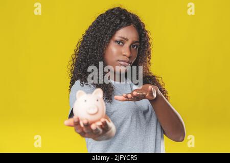 charmante fille afro-américaine aux cheveux foncés avec une banque de porc rose dans ses mains sur fond jaune Banque D'Images