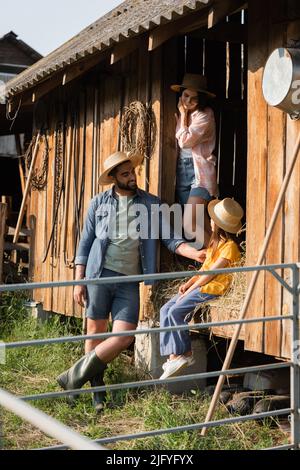 fille en chapeau de paille assise sur le foin dans la grange près de parents heureux Banque D'Images