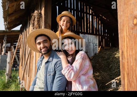 famille de village souriante dans des chapeaux de paille regardant la caméra près de la grange en bois sur la ferme Banque D'Images