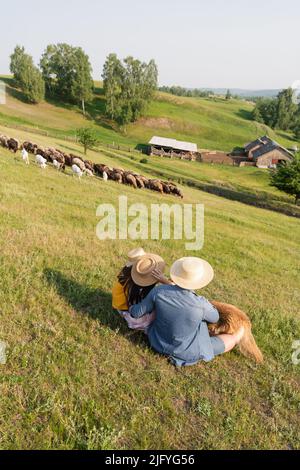 vue arrière de la famille dans des chapeaux de paille assis près du troupeau paître sur un pâturage pittoresque Banque D'Images