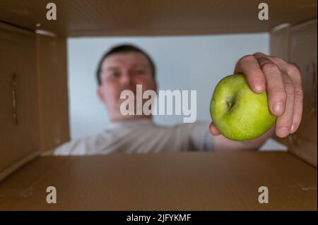 Un homme prend de la nourriture dans une boîte en carton. Une pomme verte est extraite de la boîte de don. Vue de dessous. Vue intérieure. Gros plan. Mise au point sélective. Banque D'Images