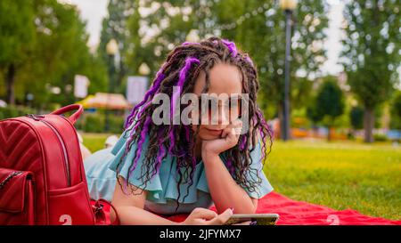 Une fille regarde une vidéo sur son téléphone, allongé sur une couverture rouge dans un parc en été. Une femme habillée regarde du contenu sur un smartphone dans un parc de la ville Banque D'Images