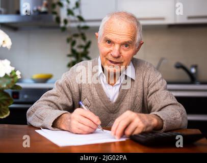 pensionné âgé écrivant sur un morceau de papier à la table dans la cuisine Banque D'Images