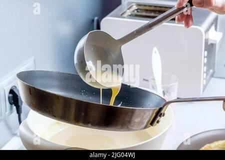 Faire couler une pâte d'un seau en métal sur une poêle graissée rouge-chaude.Le processus de cuisson des crêpes.Un régal traditionnel pour les vacances de Maslenitsa. Banque D'Images