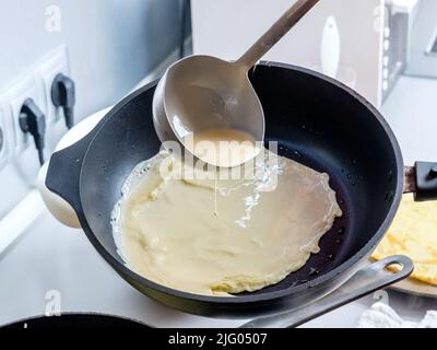 Faire couler une pâte d'un seau en métal sur une poêle graissée rouge-chaude.Le processus de cuisson des crêpes.Un régal traditionnel pour les vacances de Maslenitsa. Banque D'Images