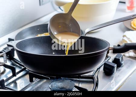 Faire couler une pâte d'un seau en métal sur une poêle graissée rouge-chaude.Le processus de cuisson des crêpes.Un régal traditionnel pour les vacances de Maslenitsa. Banque D'Images