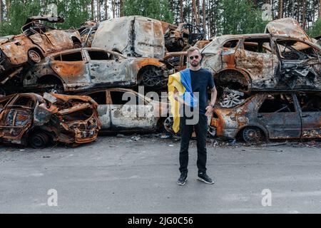 Un homme avec un drapeau ukrainien se tient contre le cimetière de voitures à Irpin près de Kiev sur beaucoup de voitures brûlées et bombardés. Conséquences des invasions militaires russes Banque D'Images