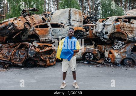 L'homme africain avec le drapeau ukrainien se tient contre le cimetière de voitures à Irpin près de Kiev sur beaucoup de voitures brûlées et bombardés. Conséquences de la milita russe Banque D'Images