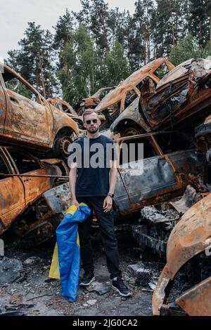 Un homme avec un drapeau ukrainien se tient contre le cimetière de voitures à Irpin près de Kiev sur beaucoup de voitures brûlées et bombardés. Conséquences des invasions militaires russes Banque D'Images