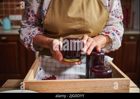 Vue rognée d'une femme dans un tablier de cuisine tenant un pot en verre de confiture de cassis sur une boîte en bois avec des pots de confiture de baies à l'envers. Collection de Banque D'Images
