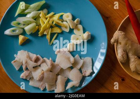 Dans l'assiette bleue, il y a du porc blanchi à l'eau chaude, des tranches d'ail et des échalotes, et du gingembre julienté; dans l'assiette en bois, il y a du poivre rouge et du gingembre. Banque D'Images