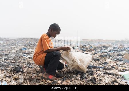 Un jeune garçon africain collecte des articles recyclables dans un grand sac en plastique à partir de tas de déchets accumulés dans une décharge qui sent les odeurs Banque D'Images