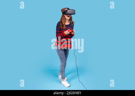 Portrait complet d'une femme portant un chandail rayé décontracté, lunettes VR, tient le joystick dans les mains, joue au jeu, expression excitée. Studio d'intérieur isolé sur fond bleu Banque D'Images