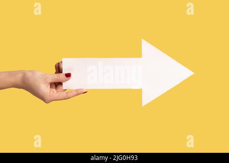 Vue de côté du profil de gros plan de la main de femme avec une flèche blanche dans les mains indiquant de côté, indiquant la direction droite. Studio d'intérieur isolé sur fond jaune. Banque D'Images