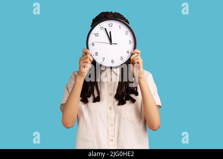 Temps de travail. Femme avec des dreadlocks se cachant face derrière une grande horloge murale, gestion du temps, rendez-vous de réunion d'affaires et de calendrier, portant une chemise blanche. Studio d'intérieur isolé sur fond bleu. Banque D'Images