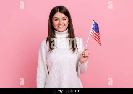 Jolie femme brune tenant le drapeau américain dans sa main et regardant la caméra avec positif, portant blanc pull de style décontracté. Studio d'intérieur isolé sur fond rose. Banque D'Images