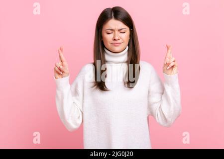 Jolie femme debout avec les yeux fermés et croisant les doigts, souhaitant bonne chance, en attente de miracle, portant blanc décontracté style pull. Studio d'intérieur isolé sur fond rose. Banque D'Images